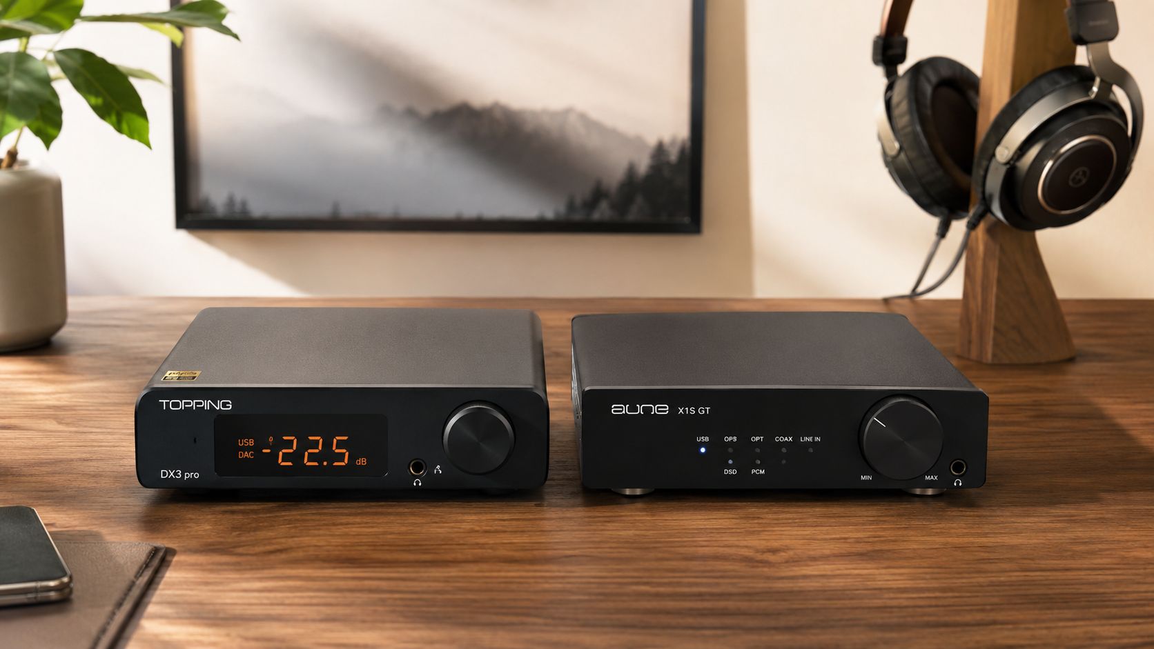 Two high-end headphone DAC and amplifier units sitting on a wooden desk with headphones in the background.