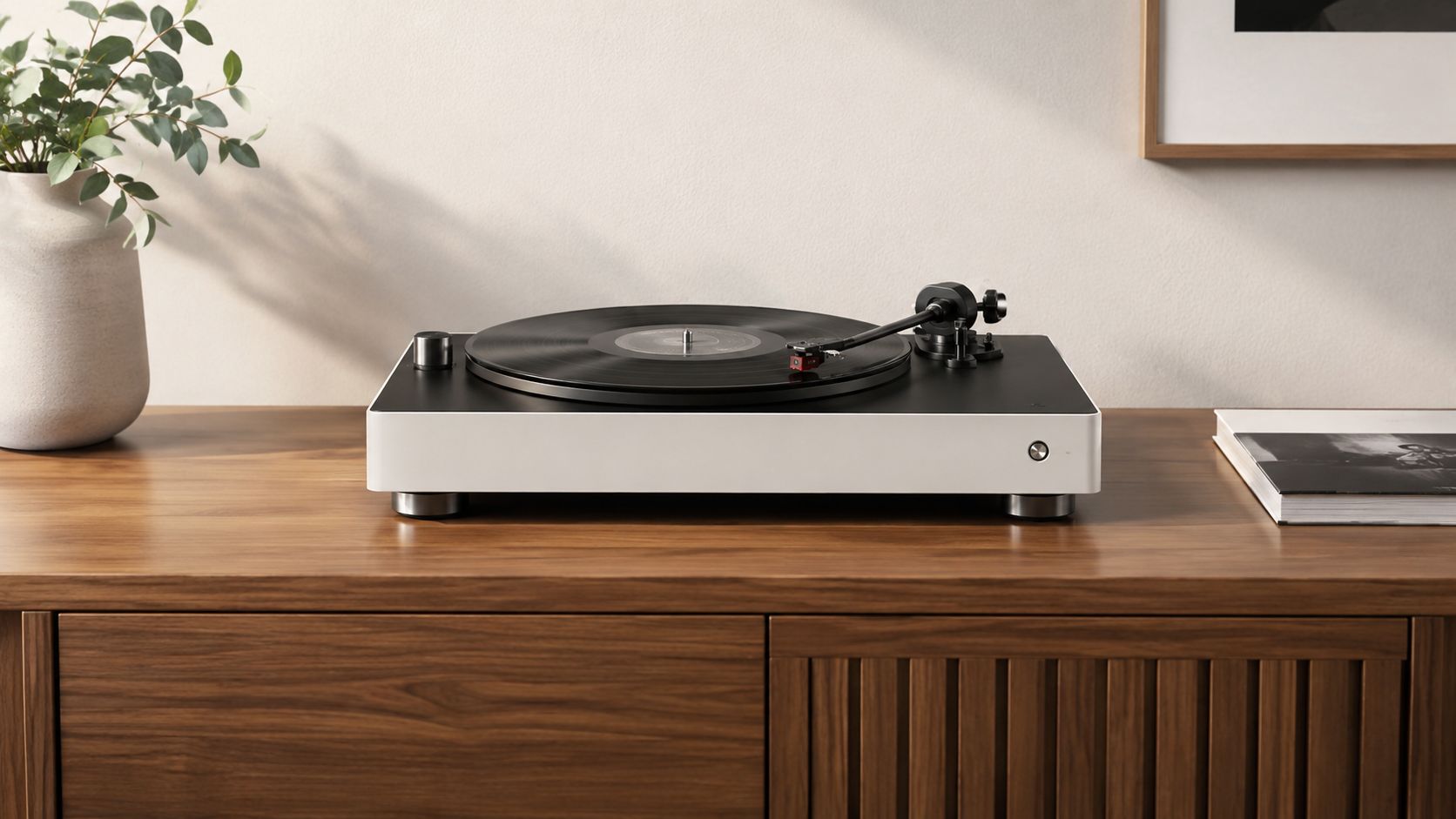 A modern, minimalist turntable sits on a wooden sideboard next to a white vase and a book.
