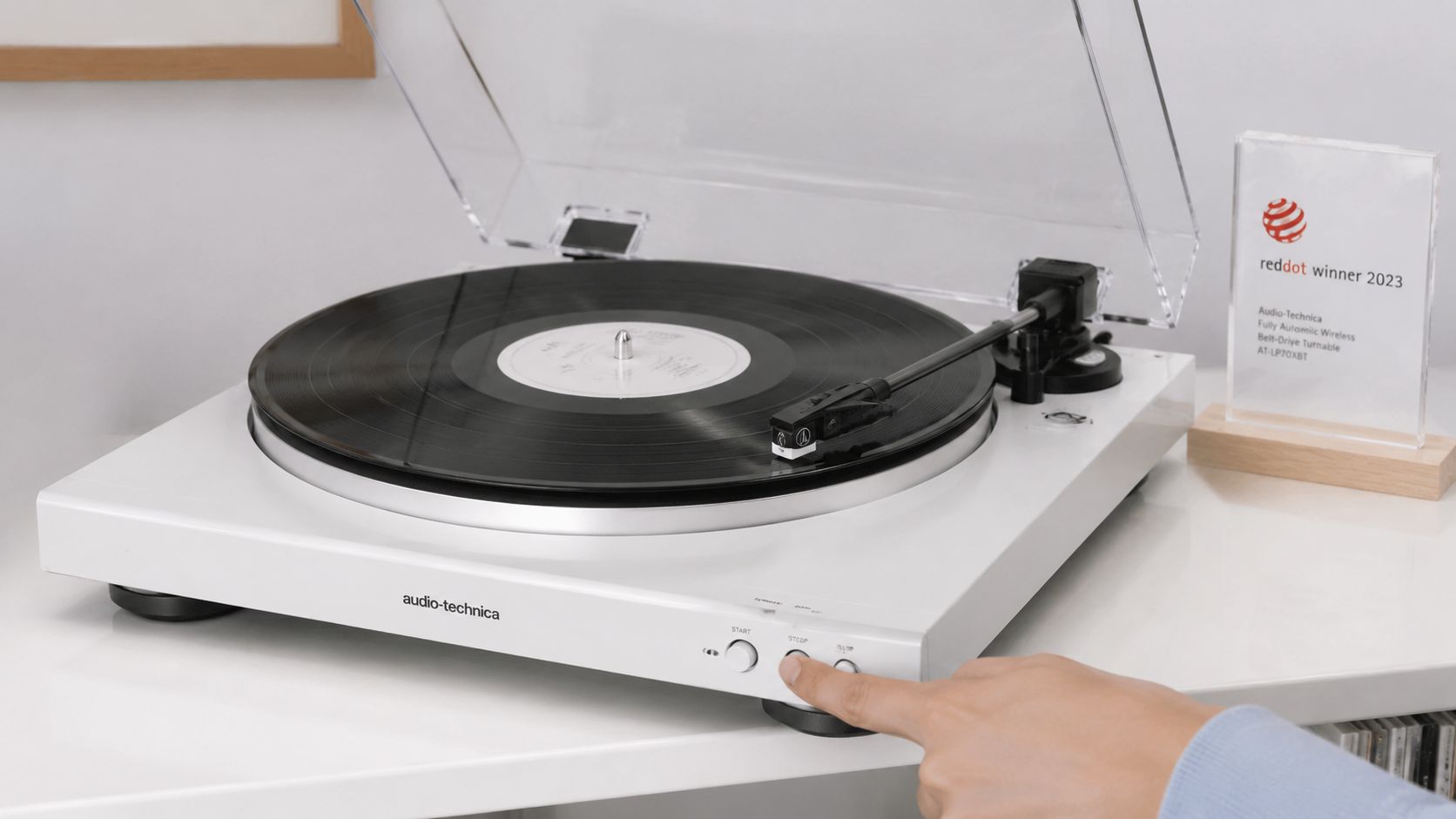 A close-up view of a white Audio-Technica turntable with a finger pressing the stop button.