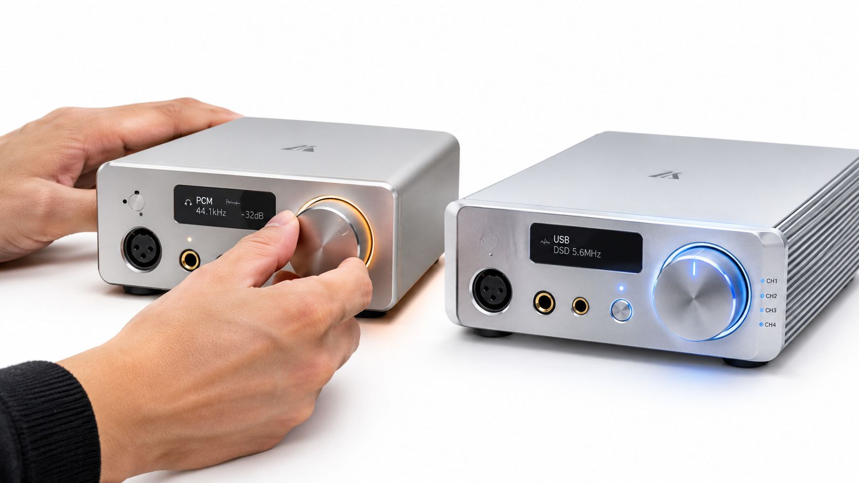 A person adjusting the volume knob on a silver headphone amplifier and DAC unit next to another identical device.